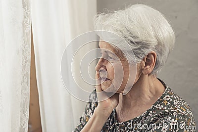 caucasian elderly pensioner lady depressed alone worried standing next to the window with white curtain Stock Photo