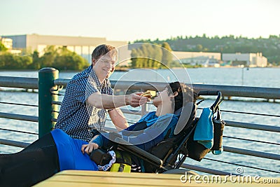 Father feeding disabled son in wheelchair hamburger by urban lake Stock Photo