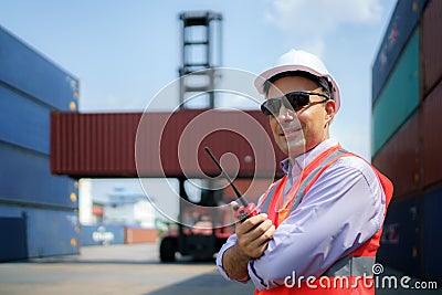 Caucasian foreman man is using walkie talkie to command his workers with crane lifting container in background in container depot Stock Photo