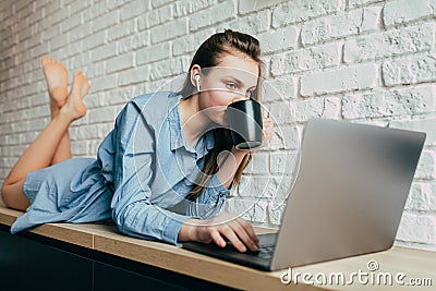 Caucasian girl in a blue shirt and long hair lies and drinks coffee while working on a computer, chatting with friends, listens to Stock Photo