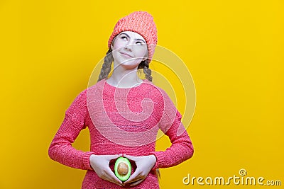 Female Fertile Concepts. Thinking Caucasian Girl In Coral Knitted Clothing Posing With Split Avocado Fruit In Front of Belly as a Stock Photo