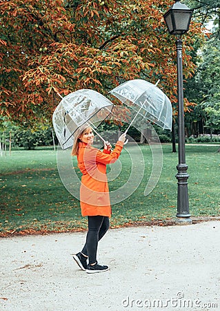 Caucasian girl in red gabardine plays with two transparent umbrellas in a park Stock Photo