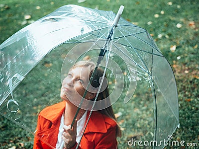 Caucasian girl with red gabardine walks through a park under a transparent umbrella Stock Photo