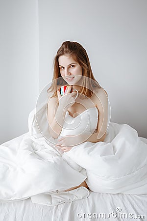 Caucasian girl woman with long red hair sitting in bed wrapped covered with blanket, holding cup Stock Photo