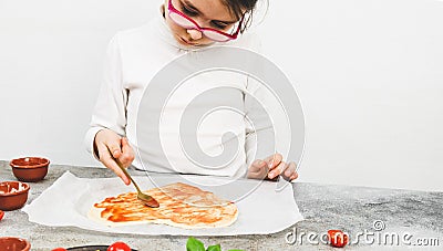 Caucasian girls in a white turtleneck smear a spoon with tomato sauce on a heart-shaped pizza dough . Stock Photo