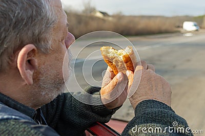 Caucasian hungry senior driver eating patty leaning his elbows on the car door Stock Photo