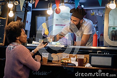 Caucasian male employee in fast food service smiling and talking to a afro-american female customer while adding mustard in her Stock Photo