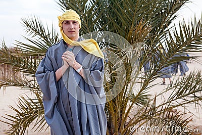 Caucasian man dressed in dishdasha and yellow headscarf standing in desert against palm tree, an European tourist Stock Photo