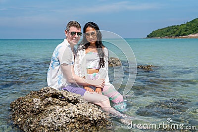 Caucasian man sit on the rock with Asian woman and look at camera with smiling on day time during honey moon vacation or holiday Stock Photo