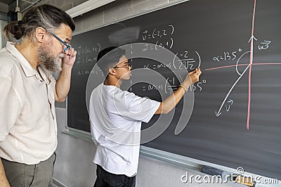 Caucasian mature man teacher supervises teen asian high school boy student writing maths exercise on blackboard with chalk Stock Photo