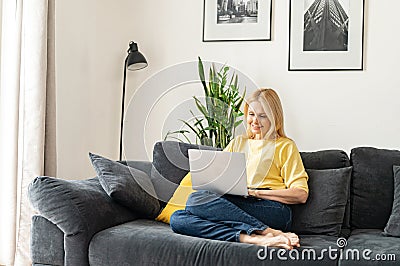 Caucasian mature woman spending home leisure with a laptop. Smiling lady sitting at the cozy sofa and looking at laptop Stock Photo