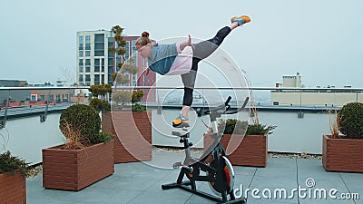 Athletic girl performing swallow acrobatic trick exercises on cycling stationary bike on house roof Stock Photo