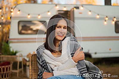 Caucasian woman sits in a wicker chair wrapped in a blanket in the yard near the trailer in autumn. Stock Photo