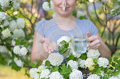 Caucasian woman takes an antihistamine medicine and removes a clothespin from her nose near a flowering tree. Stock Photo
