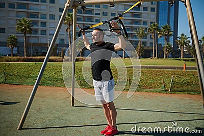 Young adult sportsman doing bodyweight exercises on arms, push ups with suspension straps in urban sports ground Stock Photo