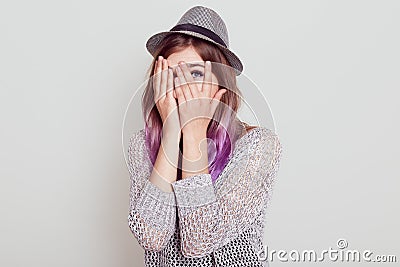 Caucasian young woman peeks through fingers, covers face with hands, dressed in hat, afraids of something, looking throw fingers Stock Photo