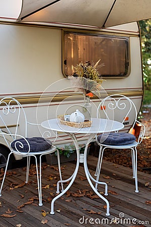 Chairs and table with tea set placed outside cozy retro caravan trailer on lawn. Interior cozy wooden RV house porch with garden f Stock Photo