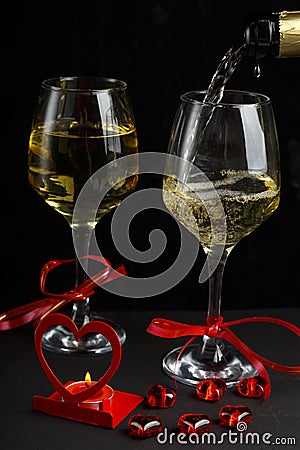 Champagne is poured into glasses on a black background next to a heart-shaped candlestick with a burning candle and a box with a Stock Photo