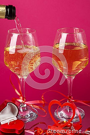 Champagne is poured into glasses on a pink background next to a heart candlestick with a burning candle and a box with a Stock Photo