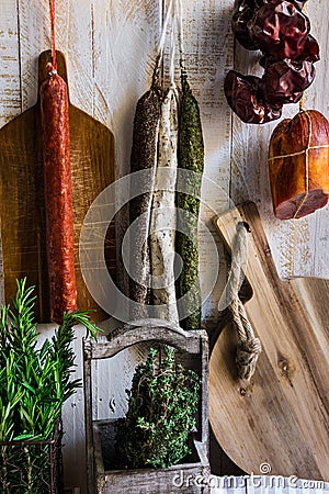 Charcuterie, variety of sausages hanging on hook, wood cutting board, string with dry peppers, fresh garden herbs, Provence Stock Photo