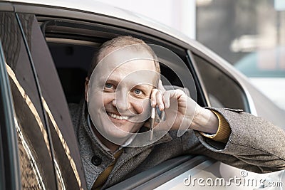 Charismatic, cheerful and handsome man with blue eyes is smiling and talking on a cell phone from an open car window. Stock Photo