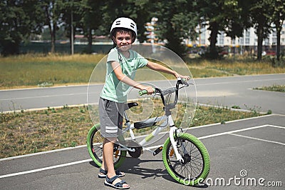 Active Caucasian child boy, in sports helmet, looking at camera, standing near his bicycle on the asphalt road outdoor. Stock Photo