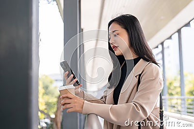 A charming Asian businesswoman chatting on her smartphone while having a quick coffee break outdoors Stock Photo