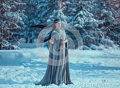 Charming attractive lady in snowy forest, militant elf princess with black long flying hair holds sword, loose gray warm Stock Photo