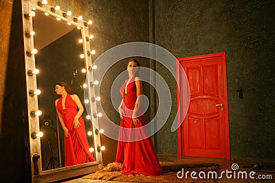 Charming beautiful girl in a red long luxury evening dress on a fur rug near a huge mirror in a frame with bulbs Stock Photo