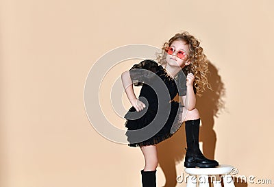 Blonde curly kid in orange sunglasses, necklace, black dress and boots. She put her foot on tabouret, posing on beige background Stock Photo