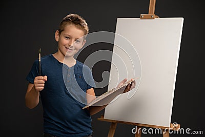 Charming boy in blue t-shirt looking at camera holding a palette with paints on the background of an easel with canvas Stock Photo