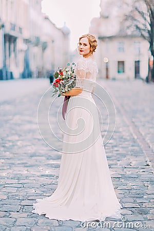 Charming bride in long lace dress holding vintage bouquet looking over shoulder into the camera with old city Stock Photo