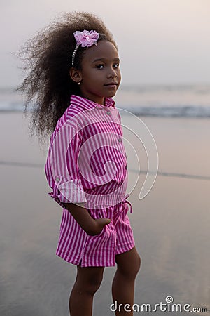 Charming dark-skinned girl with beautiful pink hair hoop looks at camera at sunset on beach. Stock Photo