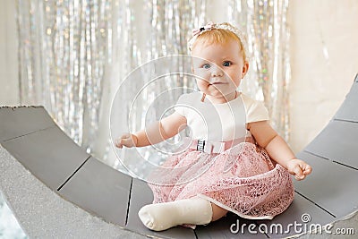 Charming little baby girl in nice pink dress, looking like a smiling angel, is sitting on the grey couch Stock Photo