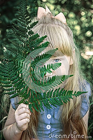 Charming little girl with blue eyes and long Golden hair is smiling, hiding behind a green fern leaf.. Happy funny tinn Stock Photo