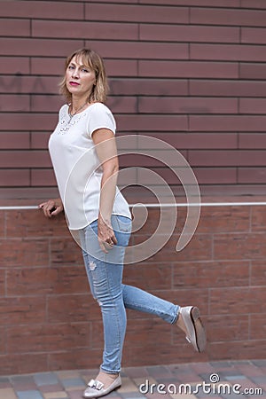 Charming mature woman, 50-55 years old, blonde, dressed in torn jeans, stands against the background of a brick wall Stock Photo