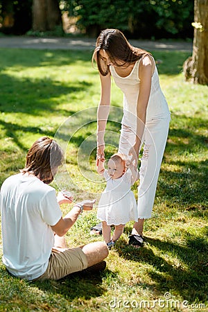 Charming mother and happy dad are teaching their little daughter wearing white dress how to make her first steps. Stock Photo