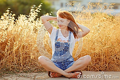 Charming red-haired girl in denim overalls sitting on a background of yellow dry grass at sunset in summer Stock Photo