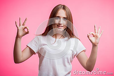 A charming red haired girl with two hands showing OK sign on pink background. Good mood, luck, happiness and emotion concept. Stock Photo