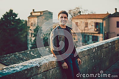 Charming smiling man outdoors with leather jacket and jeans. Leaning on a low wall. Old houses of a village in the background. Stock Photo