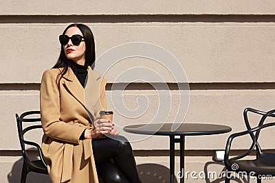 Charming stylish model with red lipstick wearing big trendy sunglasses, beige coat, black sweater and trousers, holding papercup Stock Photo
