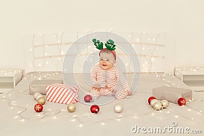 Charming toddler girl sitting on Christmas decorations bed and wearing striped long sleeve baby sleeper and festive deer horns, Stock Photo