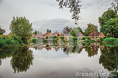 Charming wide canal next to rustic houses with green bushes and trees reflected on the water in cloudy day at Drimmelen. Stock Photo