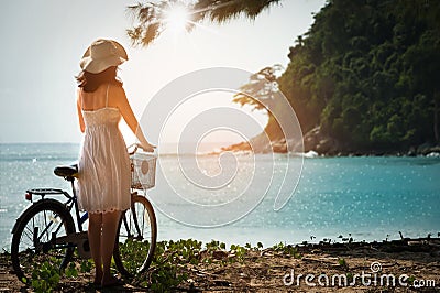 Charming woman in white dress walking bicycle enjoying blue sea. Stock Photo