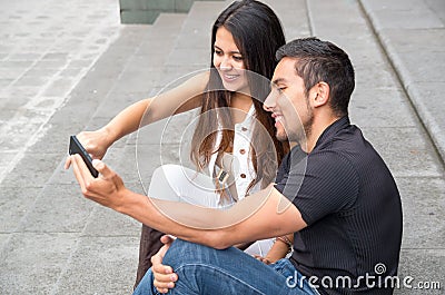 Charming young couple sitting on building steps using mobile phone and interacting happily, urban tourist concept Stock Photo