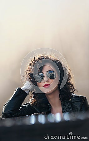 Charming young curly brunette woman with sunglasses and black leather jacket against wall. gorgeous young woman Stock Photo