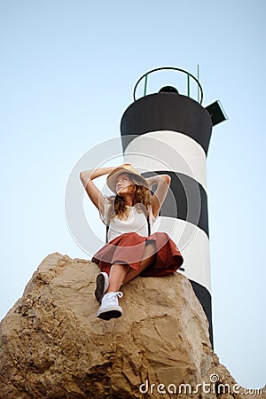 Charming young tourist woman sitting on the the large rock on background of lighthouse. Attractive ginger girl traveling by Stock Photo