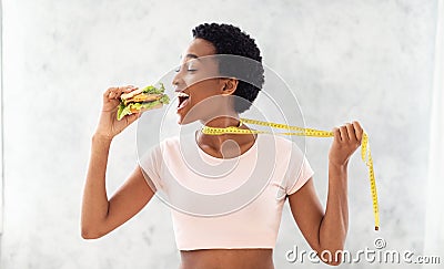 Diet breakdown. Black lady trying to eat unhealthy burger, pulling herself away by measuring tape, grey background Stock Photo