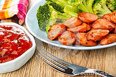 Napkin, bowl with ketchup, plate with fried sausages and cooked broccoli, fork on wooden table Stock Photo