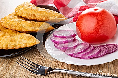 Napkin, fried cheburek in brown dish, plate with tomato and slices of red onion, fork on wooden table Stock Photo